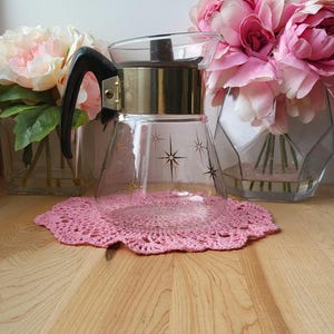 May include: A vintage glass coffee pot with a black handle and gold trim. The pot has a starburst design and is sitting on a pink crocheted doily.