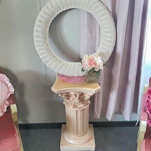 Könnte beinhalten: Ein weißer Weidenkranz sitzt auf einem Sockel mit einem rosa Blumenarrangement in einer Glasvase. Der Sockel ist aus Holz und hat eine dekorative Basis.