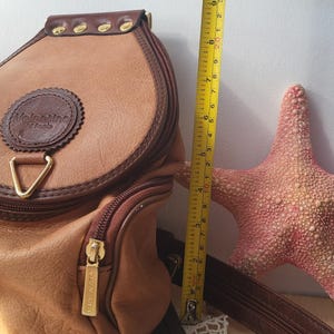 May include: A brown leather backpack with a gold zipper and a brown leather strap. The backpack has a circular logo that says "Valentino Di Renta".