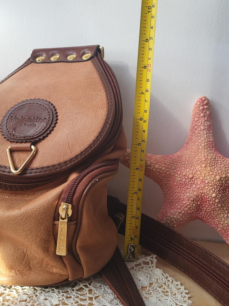 May include: A brown leather backpack with a gold zipper and a brown leather strap. The backpack has a circular logo that says "Valentino Di Renta".