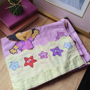 Puede incluir: Juego de sábanas rosa y amarillo con el tema de Dora la Exploradora. La sábana superior presenta a Dora saludando, con la frase "Hola amiga estrella!" y gráficos de estrellas. La sábana inferior tiene un borde amarillo con gráficos de estrellas.