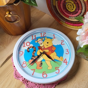 Op de afbeelding: Een witte Winnie de Poeh en Tigger klok met blauwe en groene accenten. De klok heeft zwarte wijzers en eronder een roze gehaakte onderzetter.