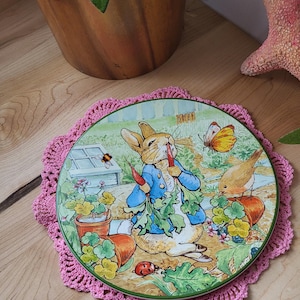 May include: A pink crocheted doily with a ceramic plate featuring a cartoon rabbit eating carrots in a garden setting. The plate has a green border and a white background with colourful flowers and a bird.