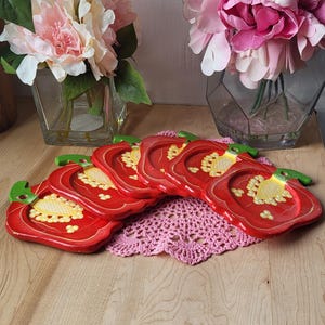 May include: Six red pepper shaped wooden coasters with green stems and yellow painted details. The coasters are arranged in a semi-circle on a pink crocheted doily and a light brown wooden surface.