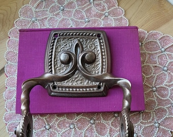 May include: Bronze-coloured metal door handle with a decorative fleur-de-lis design, mounted on a rectangular purple surface. The handle has a curved design and is set against a pink beaded placemat.