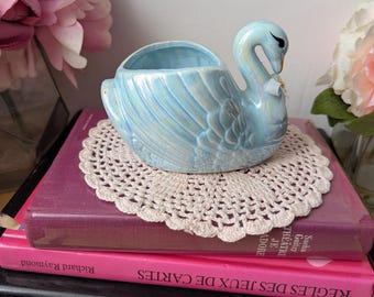 May include: A light blue ceramic swan planter with a white ribbon around its neck. The planter is sitting on a white crocheted doily on top of a stack of books.