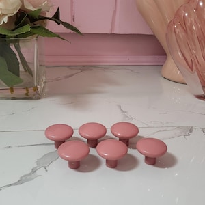 Op de afbeelding: Zeven roze kastknoppen met een afgeronde bovenkant en een korte steel, geplaatst op een wit oppervlak met marmerpatroon. Op de achtergrond staan roze decoratieve items en een vaas met bloemen. Geschikt voor meubels.