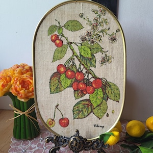 May include: Oval framed artwork featuring a cherry branch with red cherries and green leaves. The artwork is set against a beige background and framed with a gold-colored metal. The word "Cherry" is written on the artwork.