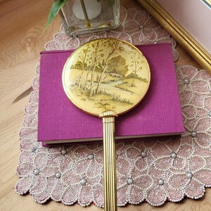 May include: A vintage hand mirror with a gold-toned handle and a round face. The mirror's face features a landscape scene in muted tones. The handle is ribbed, and the mirror rests on a purple book.