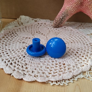 May include: Two blue plastic knobs with a round top and a short stem. The knobs are on a white crocheted doily.
