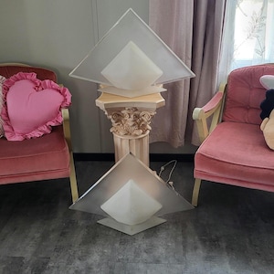 May include: Two white pyramid-shaped lamps with frosted glass shades sit on a decorative column pedestal. The lamps are on a grey floor in a room with pink chairs.