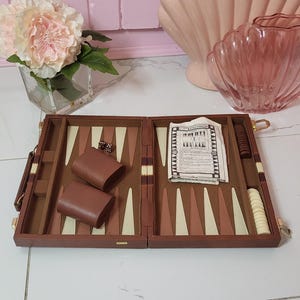 May include: Open backgammon set with brown and beige triangles, dice, and leather dice cups. The wooden case is open, revealing the playing surface and accessories. A classic board game for entertainment.