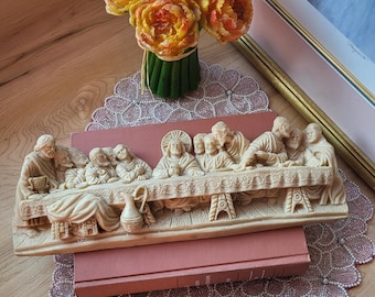 May include: A detailed, cream-colored relief sculpture depicting the Last Supper, resting on two books. The sculpture is placed on a decorative, pink doily. A bouquet of orange and yellow flowers sits nearby.