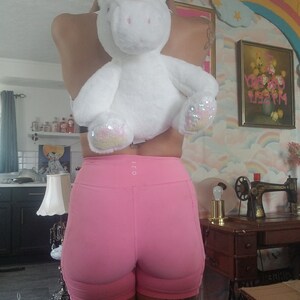 May include: A person wearing pink athletic shorts with a white unicorn plush toy strapped to their back. The unicorn has a gold horn and pink mane. The person has a tattoo on their leg that says "Maybe".