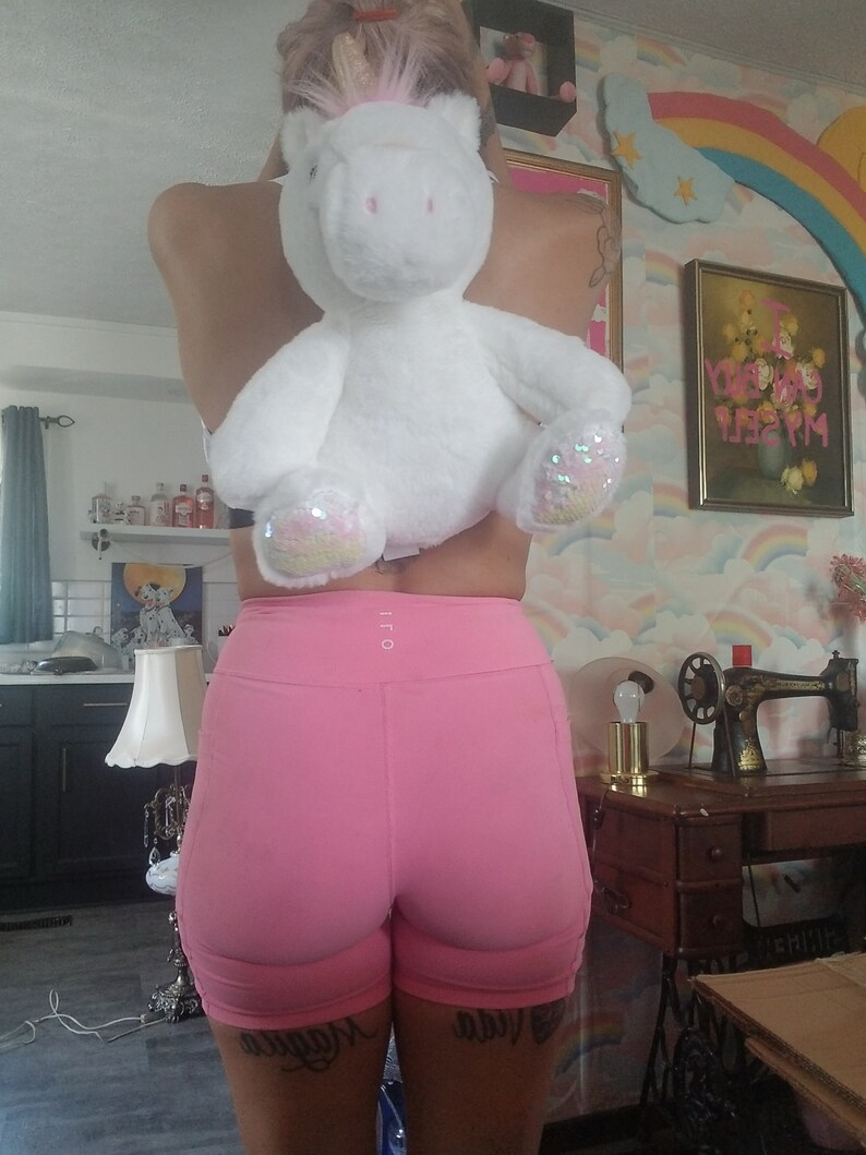 May include: A person wearing pink athletic shorts with a white unicorn plush toy strapped to their back. The unicorn has a gold horn and pink mane. The person has a tattoo on their leg that says "Maybe".