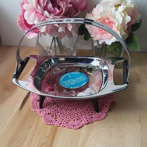 May include: Silver candle warmer with a black handle and three black legs. The warmer is sitting on a pink crocheted doily. The warmer has a blue label that says "Corning Ware 6 Hour Candle".