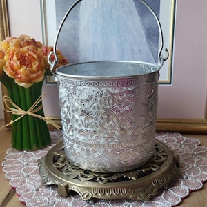 Peut inclure: Seau en métal argenté avec un motif floral en relief et une poignée. Le seau est posé sur un dessous-de-plat décoratif en laiton, présenté sur une table avec un napperon perlé rose et un bouquet de fleurs orange.