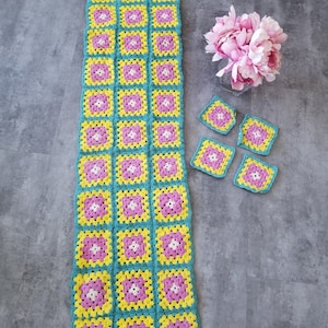 Peut inclure: Un long chemin de table crocheté en turquoise, jaune et rose avec un motif floral. Il y a aussi quatre petits carrés crochetés dans les mêmes couleurs.
