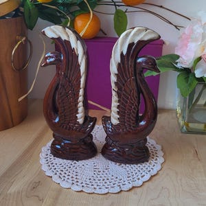 May include: A pair of brown ceramic swan figurines with white accents. The swans are standing on a white crocheted doily.