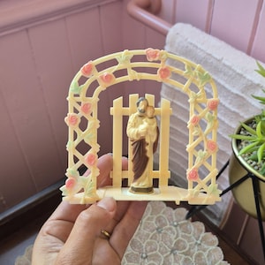 May include: A cream-colored plastic figurine of a bearded figure holding a child, positioned in front of a picket fence and beneath an arched trellis decorated with small pink roses and green leaves, all mounted on a flat rectangular base.