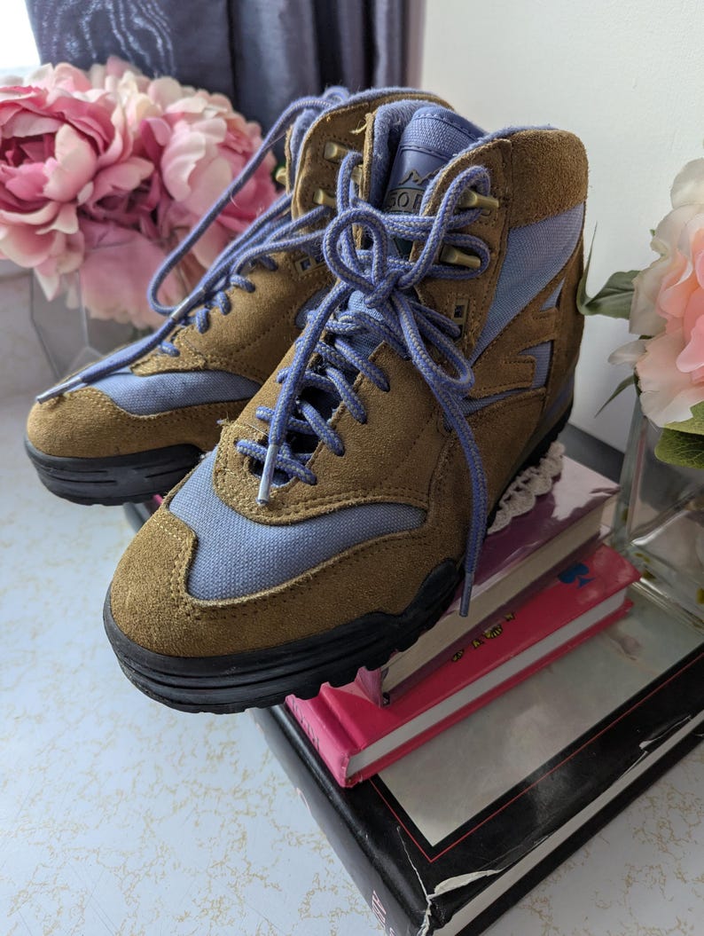 May include: Brown and blue suede hiking boots with a lace-up closure. The boots have a black rubber sole and are sitting on top of a stack of books.