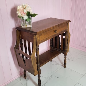 May include: A dark wooden side table with a drawer and magazine rack. The table has decorative legs and a small shelf. A glass vase with pink flowers sits on top. The table is on a white tiled floor.