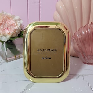 Puede incluir: Un marco de fotos rectangular dorado hecho de latón macizo. El marco tiene esquinas redondeadas y un acabado pulido. El texto en el marco dice "SOLID BRASS" y "3" x 7" (13cm x 18cm) by Bowon."