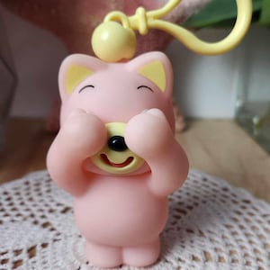 Puede incluir: Un llavero de peluche rosa y amarillo con forma de oso con las patas cubri&eacute;ndole los ojos. El oso sonr&iacute;e y tiene la nariz y la boca amarillas.