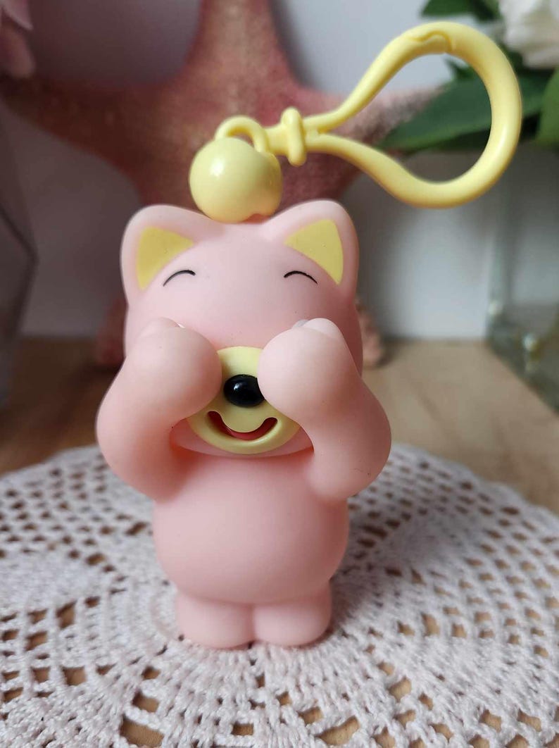 Puede incluir: Un llavero de peluche rosa y amarillo con forma de oso con las patas cubri&eacute;ndole los ojos. El oso sonr&iacute;e y tiene la nariz y la boca amarillas.
