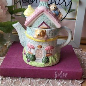 May include: A whimsical ceramic teapot shaped like a house, featuring a pink roof, light blue walls, and a yellow trim. The teapot has a handle and a spout, with teddy bear figurines on the roof and peeking from a window. It sits on a purple book.