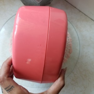 May include: A pink plastic container with a rounded shape and a smooth surface. The container is closed and has a slight seam running around the middle.