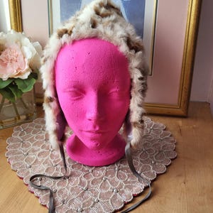 Könnte beinhalten: Ein Kunstfellhut mit braunem und cremefarbenem Muster, präsentiert auf einem pinkfarbenen Schaufensterkopf. Der Hut hat Ohrenklappen und graue Bänder. Der Hintergrund umfasst ein gerahmtes Bild und eine dekorative Tischdecke.