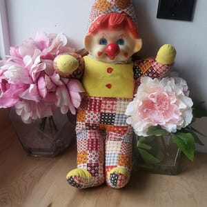May include: A vintage patchwork clown doll with red hair, a yellow shirt, and a colorful patchwork hat and pants. The doll is sitting on a wooden surface with pink flowers in the background.