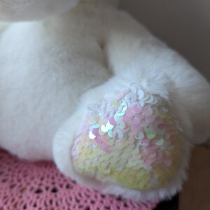 May include: White plush toy with iridescent sequins on one foot. The toy is sitting on a pink crocheted surface.