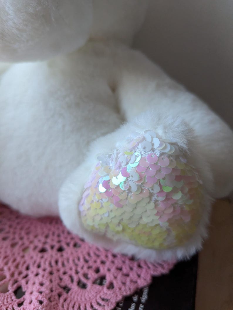 May include: White plush toy with iridescent sequins on one foot. The toy is sitting on a pink crocheted surface.