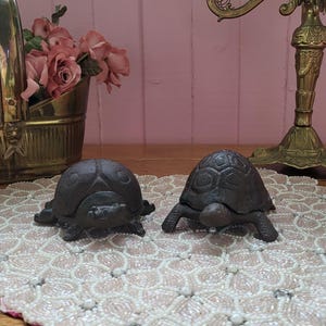 May include: Two dark brown turtle figurines with detailed shell patterns sit on a beaded placemat. The turtles are positioned in front of a brass candle holder and a decorative brass bucket with pink roses.
