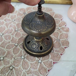 Puede incluir: Una campana de lat&oacute;n antiguo con un dise&ntilde;o floral. La campana tiene una base redonda con detalles recortados y una parte superior en forma de c&uacute;pula. La campana est&aacute; siendo presionada por un dedo. La campana est&aacute; sobre un mantel individual con cuentas.
