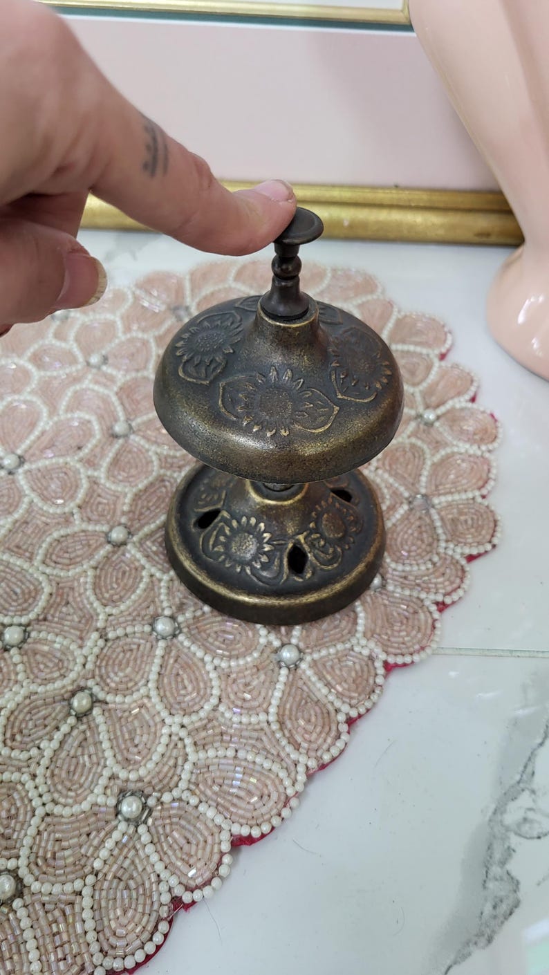 Puede incluir: Una campana de lat&oacute;n antiguo con un dise&ntilde;o floral. La campana tiene una base redonda con detalles recortados y una parte superior en forma de c&uacute;pula. La campana est&aacute; siendo presionada por un dedo. La campana est&aacute; sobre un mantel individual con cuentas.