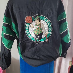Puede incluir: Chaqueta negra y verde de la marca Starter con un logotipo de los Boston Celtics en la espalda. El logotipo presenta un duende de dibujos animados sosteniendo un balón de baloncesto y un bastón. El texto "Boston Celtics" está escrito alrededor del logotipo.