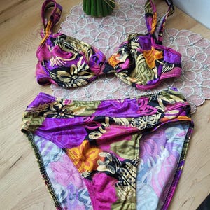 Op de afbeelding: Een tweedelige bikini met een levendig bloemenpatroon in paars, roze, geel en groen. De top heeft een klassieke driehoekvorm en de broek is een laag model. De stof heeft een zijdeachtige glans.