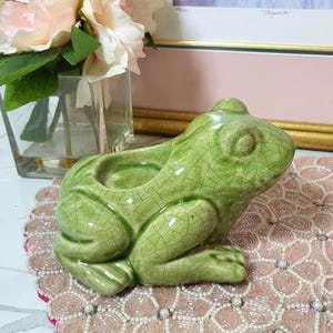 May include: A green ceramic frog-shaped planter with a cracked glaze finish. The frog is sitting on a pink beaded placemat. In the background are pink flowers in a glass vase and a framed painting.