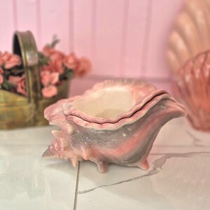 May include: A ceramic conch shell bowl featuring a gradient of soft pink and muted gray tones. The exterior displays a textured, spiky surface characteristic of a seashell, while the interior is finished in a smooth, off-white glaze.