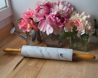 May include: A white marble rolling pin with wooden handles sits on a wooden surface.  Pink and white flowers are in glass vases behind the rolling pin.