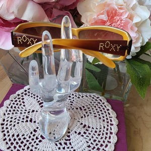 May include: Gold-toned sunglasses with brown lenses and the word "ROXY" in gold lettering on each arm. The sunglasses are displayed on a clear glass hand stand.