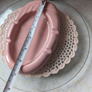 May include: A pink ceramic ashtray with a rounded oval shape and a slightly raised rim. The ashtray is approximately 6 inches long.