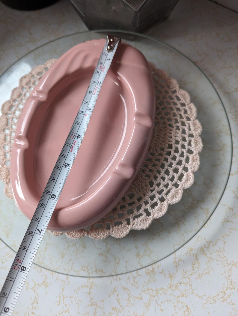 May include: A pink ceramic ashtray with a rounded oval shape and a slightly raised rim. The ashtray is approximately 6 inches long.