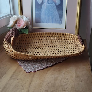 May include: A woven wicker basket with a curved design, featuring a rectangular shape and brown accents on the ends. The basket sits on a decorative doily and a wooden surface, with a framed painting in the background.