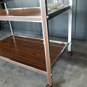 May include: A two-tiered serving cart with a brown wood top and chrome frame. The cart has four wheels and is on a gray tiled floor.