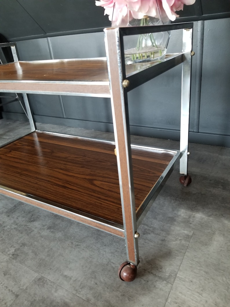 May include: A two-tiered serving cart with a brown wood top and chrome frame. The cart has four wheels and is on a gray tiled floor.