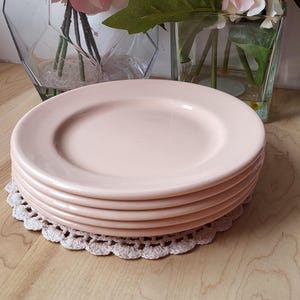 May include: A stack of five pale pink ceramic plates with a slightly raised rim. The plates are arranged on a white crocheted doily on a light wood surface.